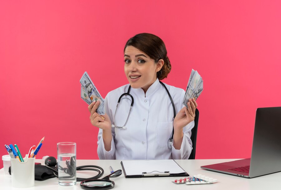 young female doctor wearing medical robe and stethoscope sitting at desk with medical tools and laptop holding money isolated on pink wall
